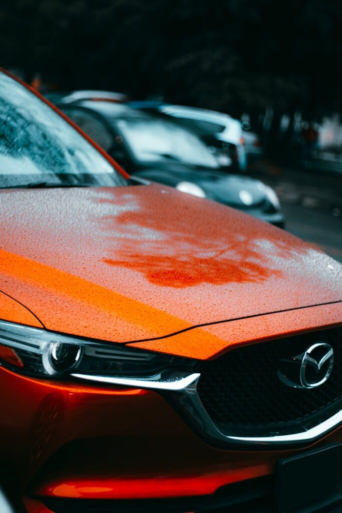 Detailed view of a red car's bonnet with raindrops, showcasing its sleek design and vibrant color in a rainy outdoor setting.